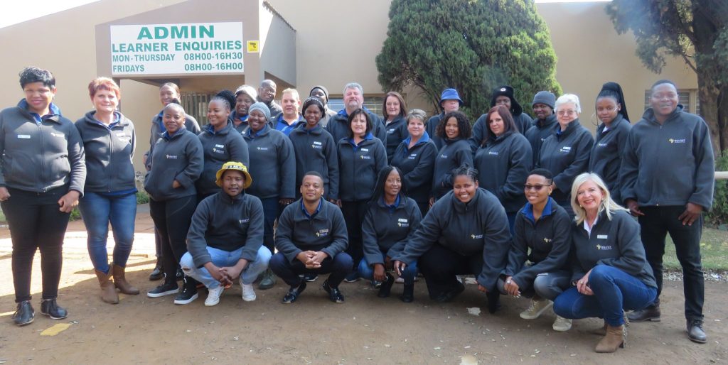 Group of individuals in matching jackets participating in learnerships, posing for a photo in front of an administration office.