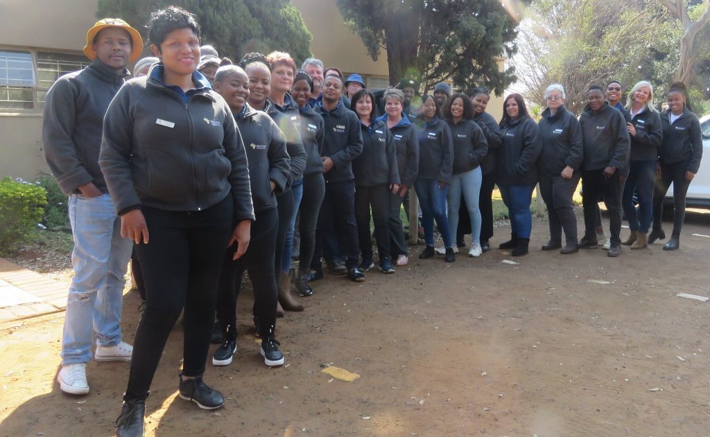 Group of smiling individuals standing outdoors wearing matching jackets, proudly displaying their certificates from completing one of the Attendance Courses designed for training people living with disabilities.