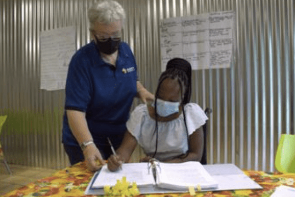 An adult in a blue shirt and mask assists a young differently-abled learner in a mask with homework at a table.