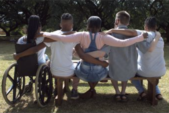 Five individuals, including people living with disabilities/differently abled learners, sitting on a bench with their backs to the camera, embracing each other around the shoulders, with one person seated in a wheelchair