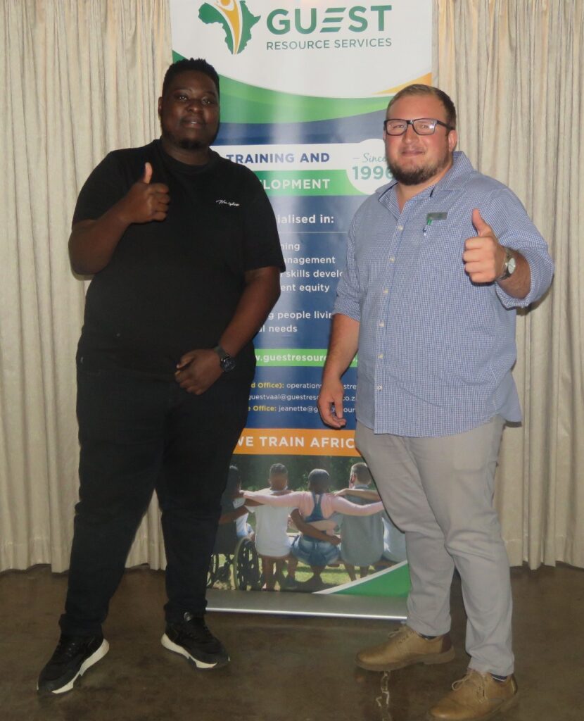 Two men standing in front of a banner, giving thumbs up, one in a black shirt and the other in a blue shirt.