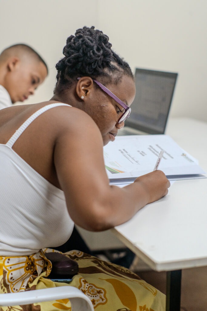 A student wearing glasses is focused on writing notes from a textbook with a laptop open in the background, unaware of the looming reality of being unemployed after graduation.