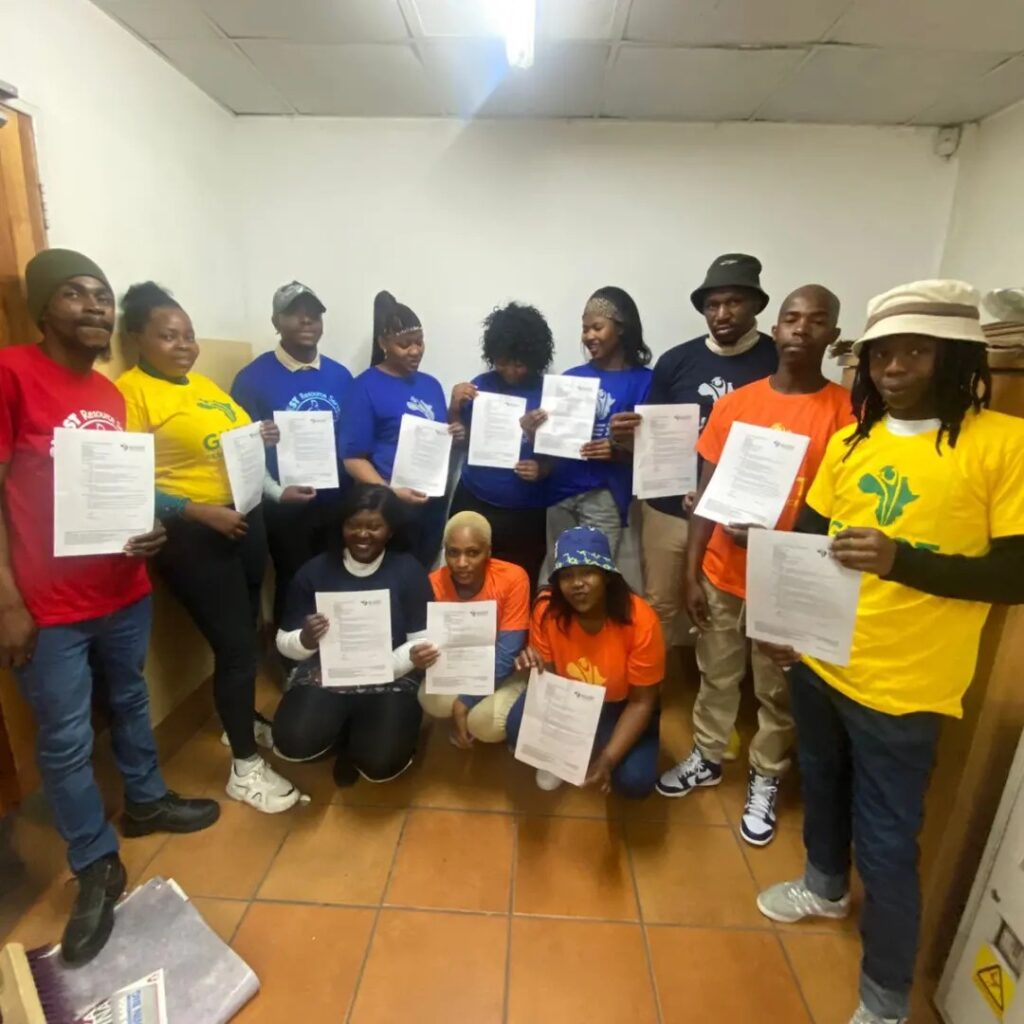 Group of people standing indoors, holding up certificates, wearing colorful shirts and hats.