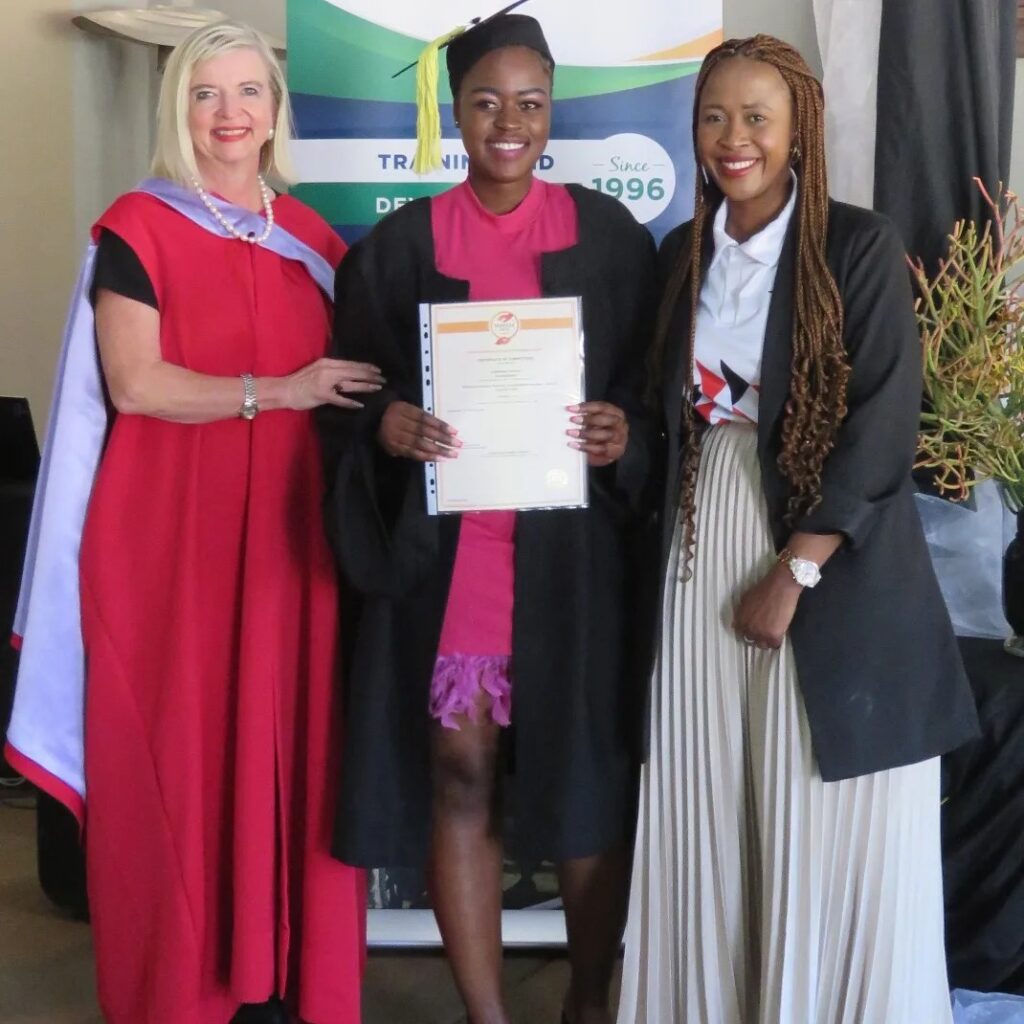 Three women pose at a graduation ceremony. One is in a red gown, another in graduation attire holding a certificate, and the third in a black coat and pleated skirt. A banner is in the background.