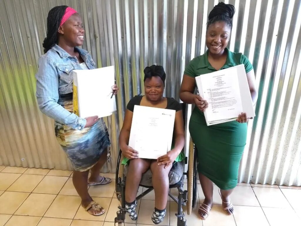Three women smiling and holding documents, with one woman in a wheelchair, standing against a corrugated metal wall.