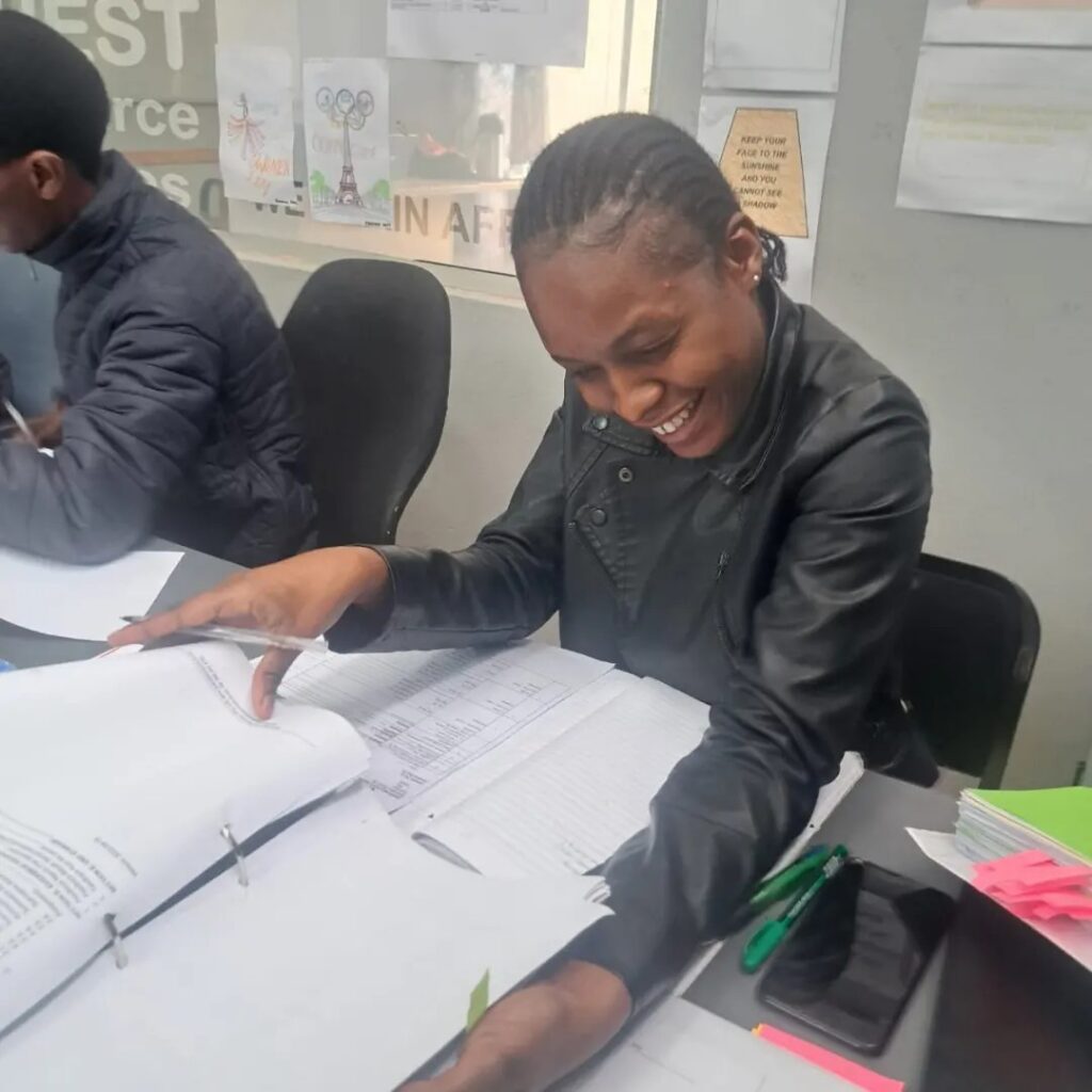 A person in a black jacket is smiling while looking at open binders on a desk. Another person is sitting beside them. Various papers are on the wall in the background.