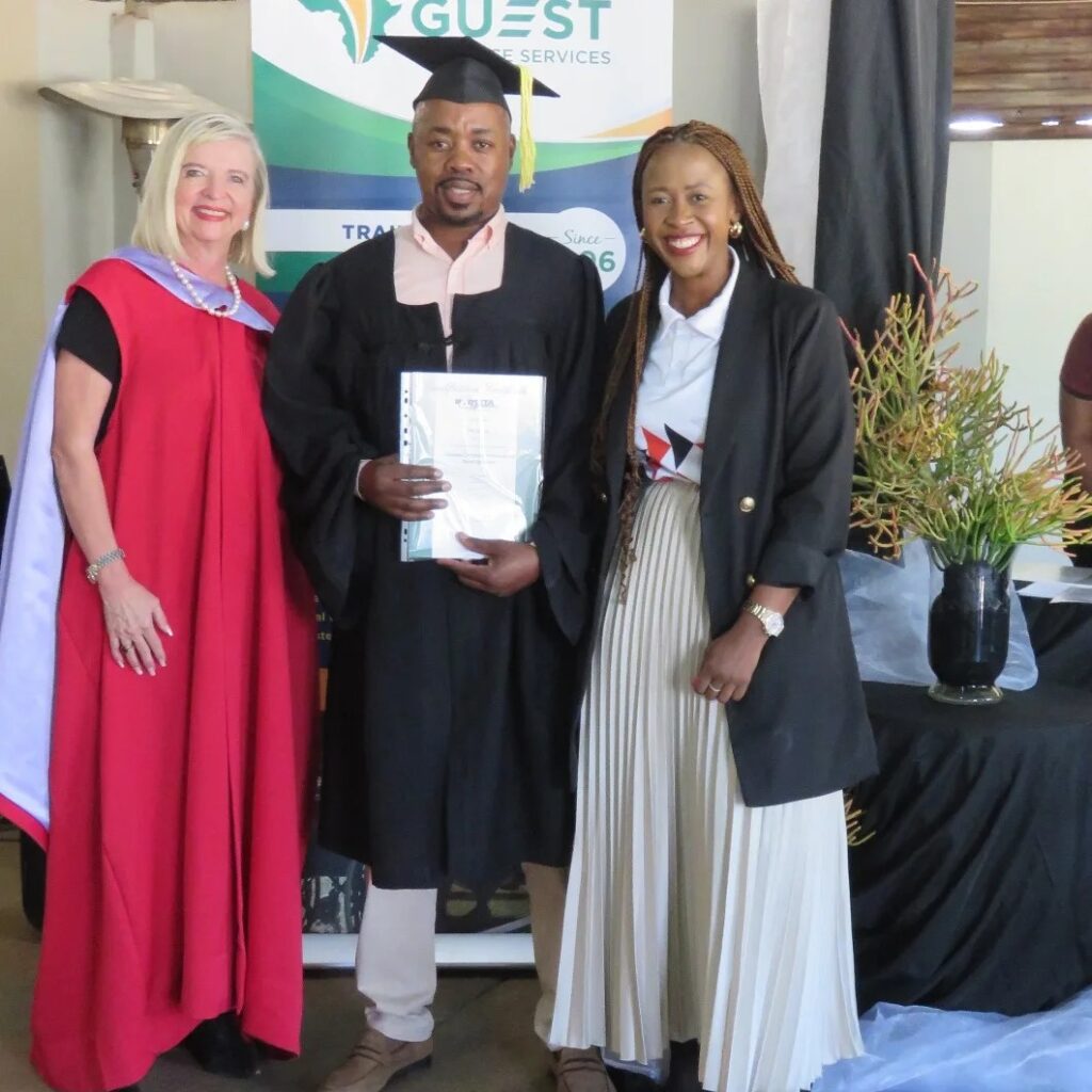 Three people stand together. The man in the center is wearing a graduation cap and gown, holding a certificate. The women on either side are smiling.