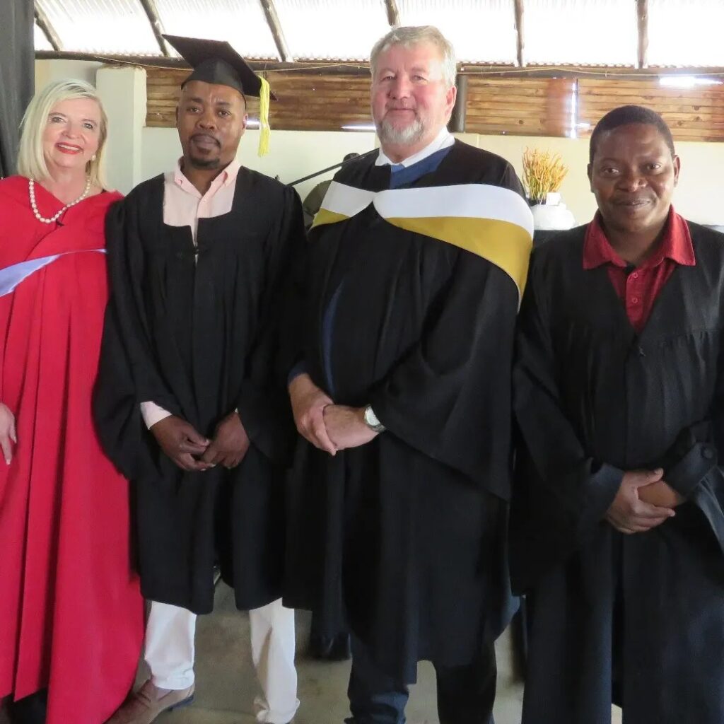 Four people in graduation attire stand indoors. The group includes three men and one woman.