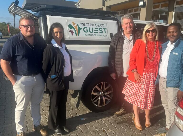 Five people stand in front of a white vehicle displaying a "We Train Africa Guest Resource Services" sign, outdoors on a sunny day.