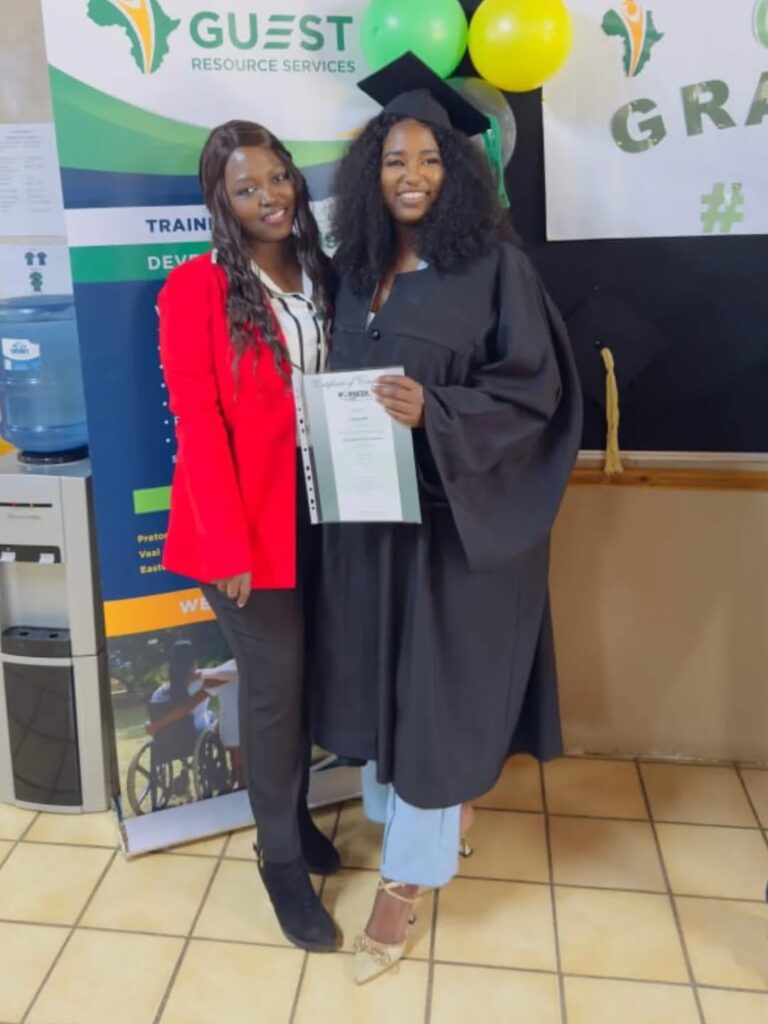 Two women pose indoors; one wears a black graduation gown and holds a certificate, while the other wears a red blazer and black pants. Graduation banner and balloons are visible in the background.