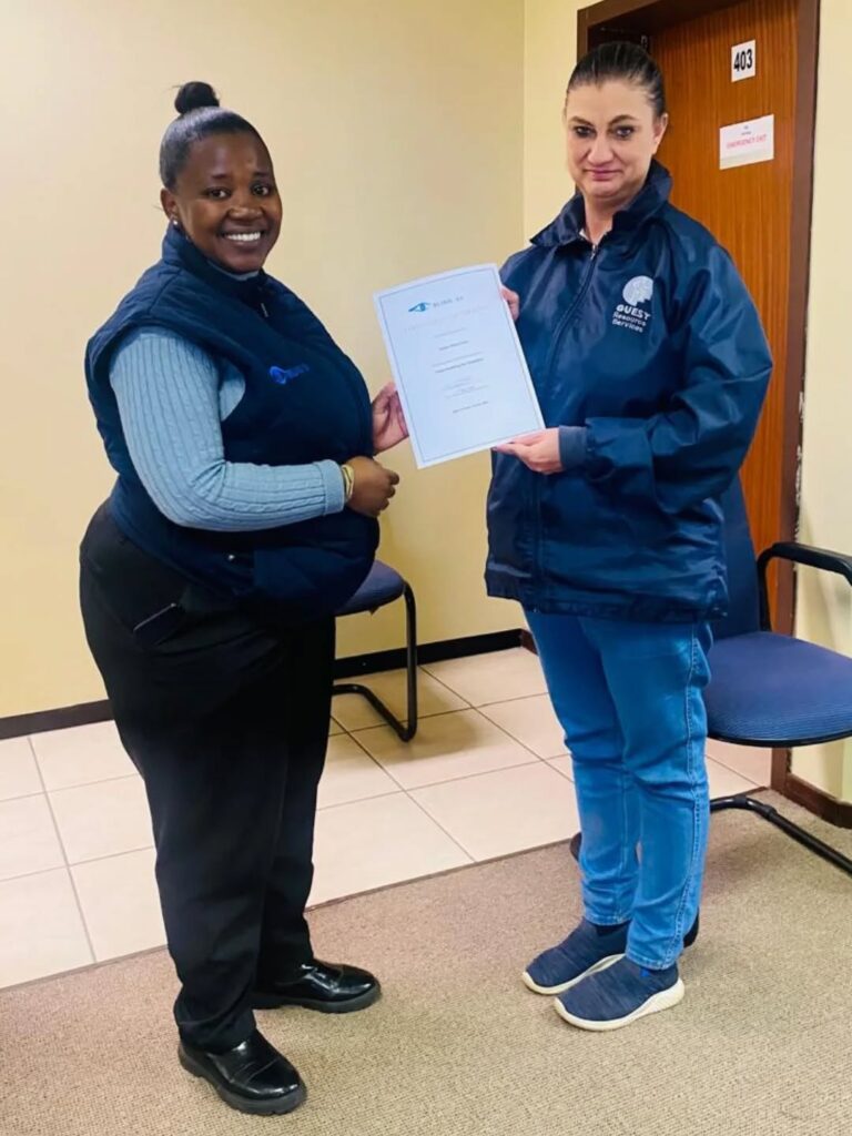 Two women stand indoors; one hands a certificate to the other. Both wear blue clothing and smile at the camera. A door labeled "403" is visible in the background.