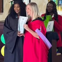 A graduate in a cap and gown stands next to an official holding a certificate, while another person stands in the background holding papers.