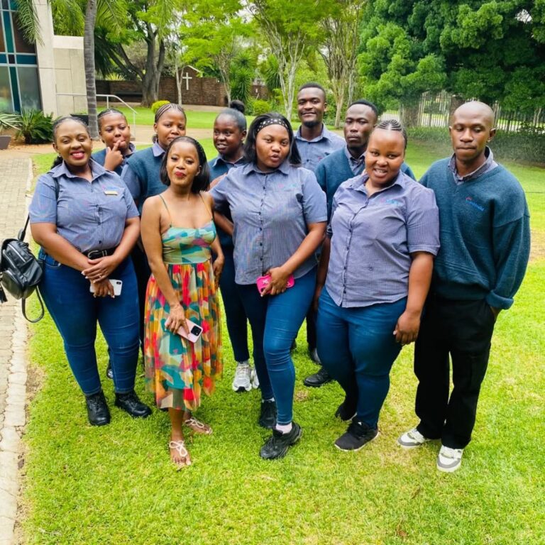 A group of ten people pose for a photo outdoors; most wear matching gray shirts and jeans, while one person in front wears a colorful dress. Trees and grass are visible in the background.