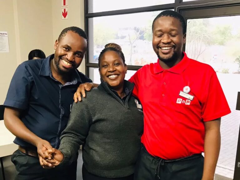 Three people stand indoors, smiling at the camera. The person on the right wears a red uniform and name tag. They appear to be in a workplace setting.