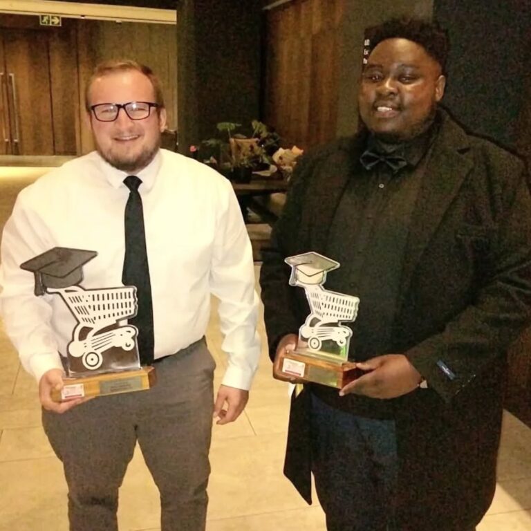 Two men in formal attire stand indoors, each holding a shopping cart-shaped trophy topped with a graduation cap.