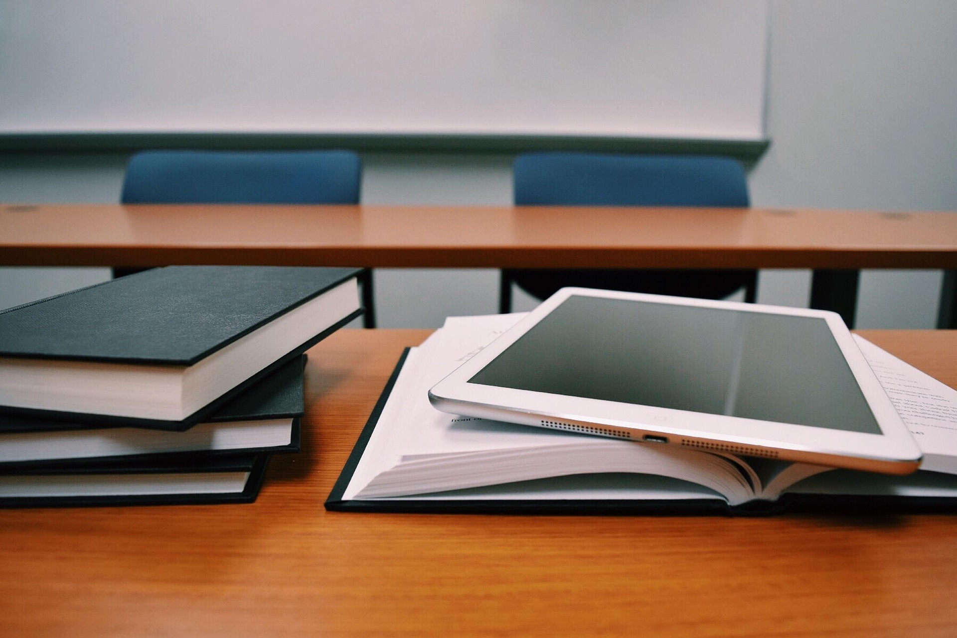 A stack of books and an open notebook with a tablet on top are on a classroom desk, with two empty chairs and a whiteboard in the background.