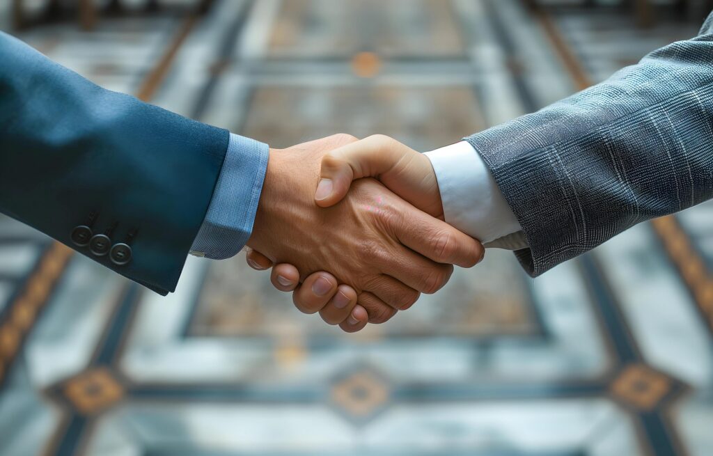 Two people in business suits shake hands indoors on a patterned tile floor, suggesting a formal agreement or partnership.