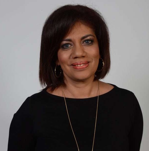 A middle-aged woman with shoulder-length dark hair wearing a black top and gold hoop earrings.