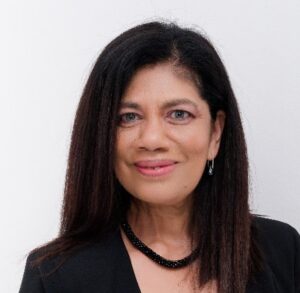 A headshot of a woman with long dark hair, wearing a black top and a beaded necklace.
